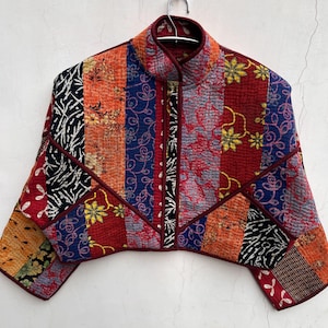 Puede incluir: Chaqueta patchwork con cuello mandarín, con una variedad de patrones de tela en rojo, naranja, azul y negro. La chaqueta tiene una silueta recortada y está colgada de una percha de metal. El diseño incluye motivos florales y abstractos.