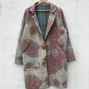Könnte beinhalten: Ein langärmeliger Mantel mit gesteppter Textur und einem floralen Muster in Rosa, Grau und Weiß. Das Kleidungsstück hat einen Reverskragen, zwei Vordertaschen mit brauner Paspelierung und zwei Knöpfe. Das Innenfutter zeigt ein blau-braunes Karomuster.
