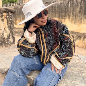 May include: A white sun hat with a wide brim, decorated with a tiger print. The person is wearing a black jacket with gold trim and blue jeans. The jacket has a colourful stitched pattern.