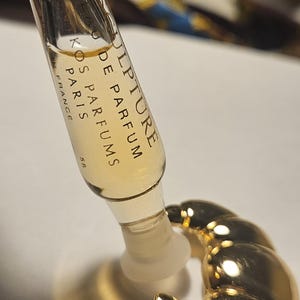 Puede incluir: Un vial de muestra de perfume con el texto "Sculpture de Parfum Kos Paris France" se sostiene sobre un soporte de perfume dorado en forma de croissant. El vial contiene un líquido ámbar claro.