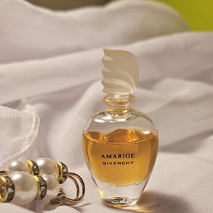May include: A small, clear glass perfume bottle with a decorative stopper, containing a golden liquid. The bottle is labelled "Amarige Givenchy." A pearl and gold bracelet is in the foreground, set against white fabric.