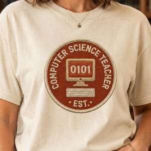 Puede incluir: Una camiseta de color crema con un gráfico circular de color óxido en el pecho. El diseño incluye un monitor de ordenador que muestra el texto 0101, un teclado y el texto COMPUTER SCIENCE TEACHER EST. dispuesto en un borde circular.