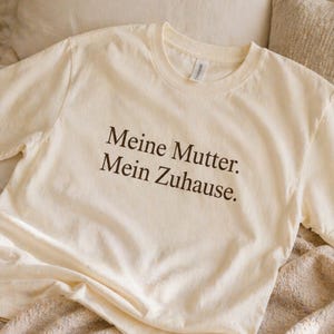 Puede incluir: Camiseta color crema con el texto en alemán "Meine Mutter. Mein Zuhause." impreso en marrón oscuro. La camiseta tiene cuello redondo y mangas cortas, y está hecha de un material suave.