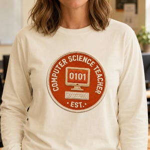 Puede incluir: Una camiseta de manga larga color crema con un parche circular bordado en el pecho. El parche es naranja con un borde blanco y muestra un monitor de computadora con el texto 0101, un teclado y las palabras COMPUTER SCIENCE TEACHER EST.