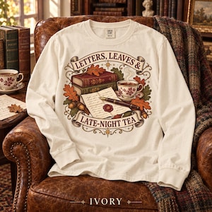 Può includere: Una maglia a maniche lunghe color avorio con una stampa grafica di libri impilati, una penna stilografica, una tazza da tè e foglie autunnali. Il testo "LETTERS, LEAVES & LATE-NIGHT TEA" è inserito in nastri decorativi con ghiande e stelle.