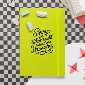 Peut inclure: Un cahier jaune vif avec le texte "Sorry for What I said when I was Hungry" en écriture noire. Le cahier est fermé et possède une bande élastique jaune. Des guimauves aux couleurs pastel sont éparpillées autour du cahier.