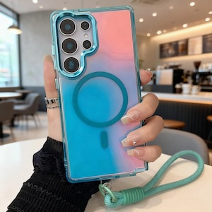 May include: A clear phone case with a gradient design of pink and blue. The case has a blue border and a circular magnetic ring. A teal lanyard is attached to the bottom. The phone case is designed for a smartphone.