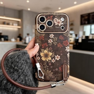 Op de afbeelding: Een bruine telefoonhoes met bloemenpatroon en een bijpassende bruine riem. De hoes heeft een gedetailleerd bloemenpatroon in geel, rood en wit. De camera heeft een goudkleurige rand. De riem is bevestigd met zilverkleurige hardware. Het woord "Flower" staat op de hoes.
