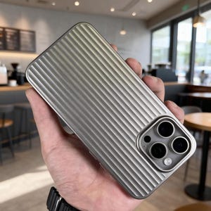 Könnte beinhalten: Silberne Handyhülle mit gerilltem Design. Die Hülle hat einen Kameraausschnitt mit drei Objektiven. Die Hülle wird in einer Hand gehalten, wodurch die metallische Oberfläche und die Kamerafunktionen des Telefons hervorgehoben werden.