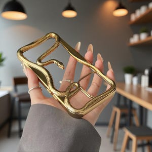 May include: A gold-coloured phone case featuring a snake design. The case has a textured surface and a stylized snake wrapping around the edges. The case is held in a hand.