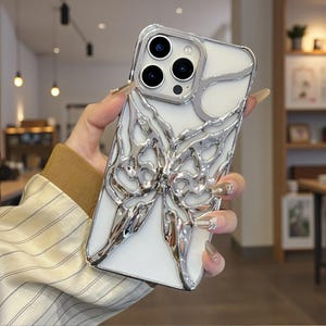 May include: A white phone case featuring a silver, abstract butterfly design. The case has a clear border and a camera cutout. The butterfly design is raised and has a metallic finish, adding a decorative element to the phone.
