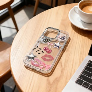 Op de afbeelding: Transparante telefoonhoes met zilveren sterren, roze kersen, lippen, strikken en het nummer 777. De hoes ligt op een houten tafel naast een kopje koffie.