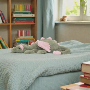 Puede incluir: Un juguete de peluche con forma de drag&oacute;n verde con alas y p&uacute;as rosas descansa sobre una colcha azul claro. El peluche est&aacute; dise&ntilde;ado con una apariencia amigable, adecuado para la habitaci&oacute;n de un ni&ntilde;o o como objeto decorativo. El drag&oacute;n mide unos 40 cm de largo.
