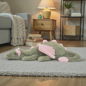 Puede incluir: Un peluche verde con forma de drag&oacute;n, con alas y detalles rosas, tumbado sobre una alfombra gris. El drag&oacute;n tiene un cuerpo largo, p&uacute;as rosas y una expresi&oacute;n amigable. Este juguete para ni&ntilde;os a&ntilde;ade un toque caprichoso a cualquier habitaci&oacute;n.