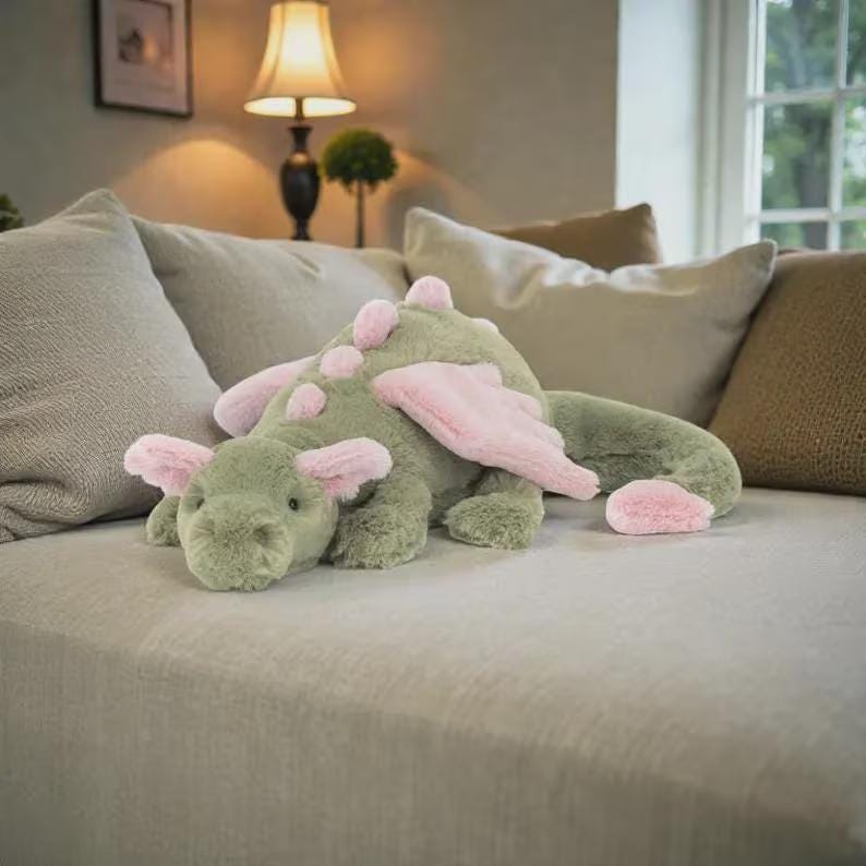 Puede incluir: Un peluche de drag&oacute;n verde con alas rosas, detalles de orejas y acentos de espina dorsal. El peluche est&aacute; tumbado en un sof&aacute; de color claro. El drag&oacute;n tiene una expresi&oacute;n amigable y est&aacute; dise&ntilde;ado para ni&ntilde;os.