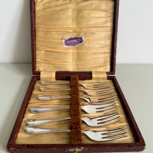 Könnte beinhalten: Ein Set aus sechs versilberten Gabeln in einer braunen, strukturierten Präsentationsbox mit goldfarbenem Satin-Innenfutter. Ein kleines violettes Etikett im Inneren trägt die Aufschrift EPNS Sterling Silver Plated Finest Finish Made in Sheffield Eng.