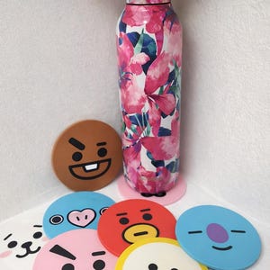 Puede incluir: Una botella de agua de acero inoxidable con un estampado floral en rosa y blanco, rodeada de siete posavasos redondos y coloridos con caras de personajes de dibujos animados. La botella mide unos 25 cm de alto.