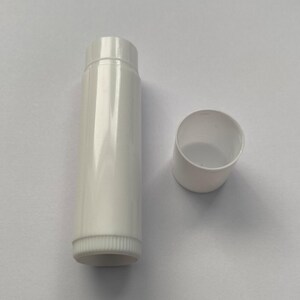 May include: A white lip balm tube and its cap. The tube is cylindrical with a ribbed base and a smooth, glossy finish. The cap is off, showing the tube's opening. The product is designed for lip care.