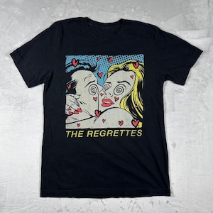 May include: Black t-shirt featuring a pop art style graphic of a couple kissing, surrounded by hearts. The graphic has a blue and yellow color scheme. The text "THE REGRETTES" is printed in yellow at the bottom of the graphic.