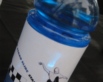 Puede incluir: Una botella de bebida deportiva azul con una etiqueta blanca que muestra a un patinador haciendo un board slide stall. La etiqueta también incluye el texto "(SK8R.ADE)".