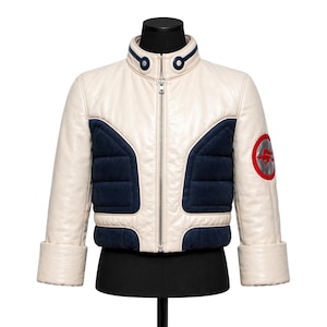May include: Cream-colored leather jacket with navy blue quilted panels and a stand-up collar. Features a silver zipper, rolled cuffs, and a red and silver emblem on the sleeve. The jacket is displayed on a black mannequin.
