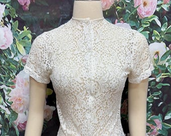 May include: A white lace top with short sleeves and a high neckline. The top has a button-down front and a split back. The lace pattern is intricate and detailed, creating a delicate and feminine look. The top is displayed on a mannequin.