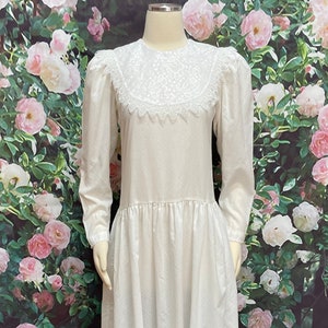 May include: A white long-sleeved dress with a lace collar and a gathered skirt. The dress is photographed against a backdrop of pink roses.