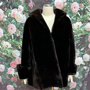 May include: A dark brown faux fur coat with a shawl collar and turned-up cuffs. The coat is shown on a mannequin against a floral backdrop of pink and white roses.