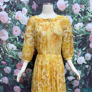 May include: A yellow floral print dress with three-quarter sleeves and a pleated skirt. The dress features a round neckline with a tie detail. The floral pattern includes white and orange accents. The dress is displayed on a mannequin.