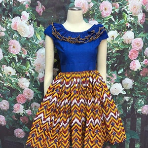 May include: A blue and yellow dress with a ruffled neckline and a geometric print skirt. The dress is on a mannequin against a backdrop of pink and white roses.