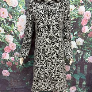 May include: A black and white patterned coat with a circle design. The coat has a collar, long sleeves, and three black buttons. The coat is displayed on a mannequin. The background features a floral design.