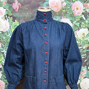 May include: A dark blue denim blouse with a high collar and long, puffed sleeves. The blouse features red buttons down the front and two front pockets. Red stitching accents the seams.