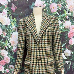 May include: A vintage blazer in a classic houndstooth pattern. The jacket features a mix of green, brown, and tan colors. It has two front pockets and a two-button closure. The blazer is displayed on a mannequin.