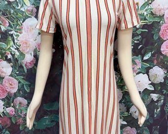 Puede incluir: Un vestido de manga corta con un patrón de rayas verticales en naranja y negro sobre un fondo color crema. La prenda tiene un cuello alto con una cremallera central que se extiende por la parte delantera, creando una silueta estructurada.