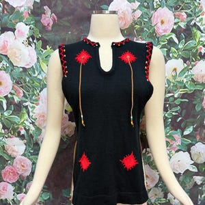 May include: Black sleeveless top with red embroidered details. The top features a V-neckline, red patches, and decorative gold-colored strings with beads. The edges are trimmed with red zig-zag stitching. The top is displayed on a mannequin.