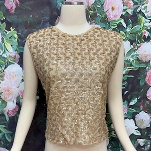 May include: A sleeveless, gold-toned top with a textured, sequined design. The top has a round neckline and a slightly gathered hem. The garment is displayed on a mannequin, showcasing its intricate details and shimmering appearance. The background features a floral pattern.