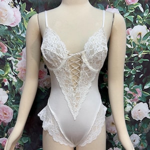 May include: White lace bodysuit with a plunging neckline and delicate spaghetti straps. The bodice features a lace-up detail and is trimmed with intricate lace. The bottom portion has a ruffled lace edge. The background is a floral pattern.