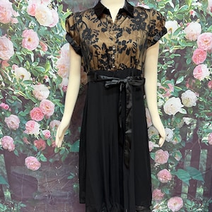 May include: A black dress with a sheer, floral-patterned bodice and a black skirt. The dress features a black satin tie belt and short sleeves. The dress is displayed on a mannequin.