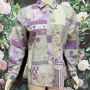 May include: A long-sleeved patchwork shirt in shades of lavender, green, and white. The shirt features a button-down front, a collar, and various patterned patches with heart and floral motifs. The shirt is displayed on a mannequin.