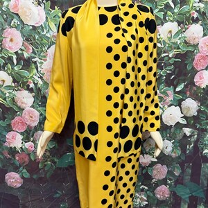 May include: A vintage yellow two-piece suit with black polka dots. The suit includes a long-sleeved jacket and a skirt. The jacket has a scarf-like collar. The suit is displayed on a mannequin.