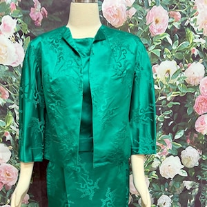May include: A green three-piece suit with a jacket, dress, and matching top. The fabric has a subtle floral pattern.