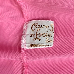 May include: A bright pink fabric garment featuring a white rectangular clothing label sewn into the seam. The label displays the gold embroidered text Claire S by Lucie Be, with a visible stitched border and a fine ribbed texture on the pink material.