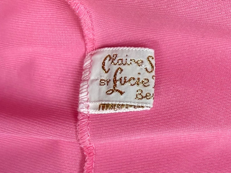 May include: A bright pink fabric garment featuring a white rectangular clothing label sewn into the seam. The label displays the gold embroidered text Claire S by Lucie Be, with a visible stitched border and a fine ribbed texture on the pink material.