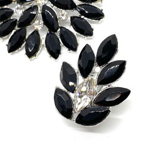 May include: Two silver brooches featuring black and clear rhinestones. The larger brooch is circular with a cluster of black rhinestones. The smaller brooch is leaf-shaped with a single clear rhinestone in the centre.