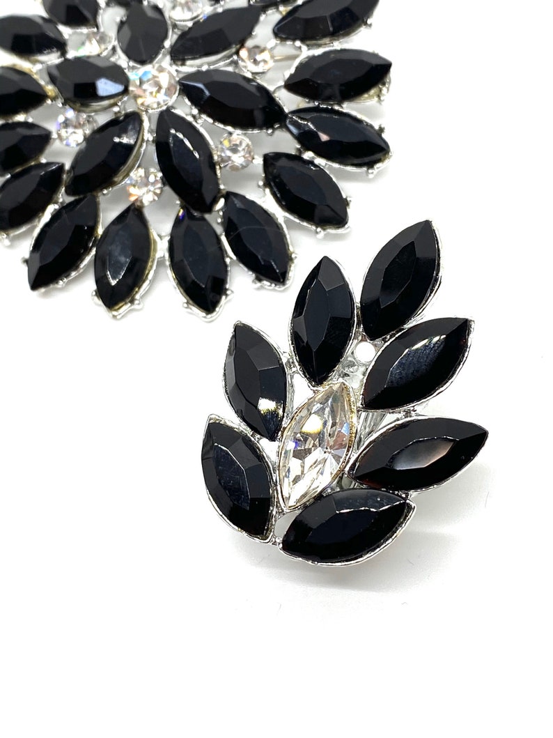 May include: Two silver brooches featuring black and clear rhinestones. The larger brooch is circular with a cluster of black rhinestones. The smaller brooch is leaf-shaped with a single clear rhinestone in the centre.