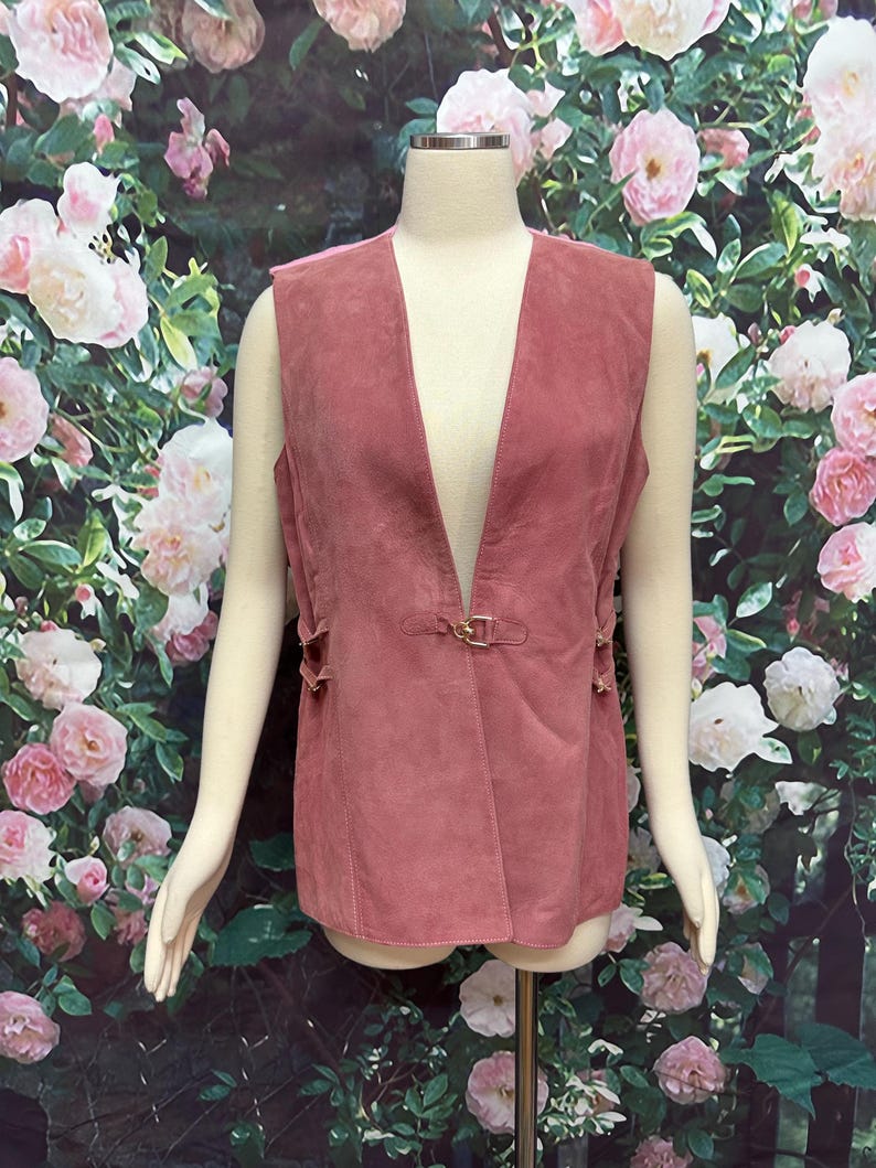 May include: A dusty rose suede vest with a deep V-neckline and a gold buckle closure. The vest has a sleeveless design and features decorative stitching along the sides.