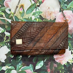 May include: A rectangular brown snakeskin-patterned clutch bag featuring diagonal panels of varying dark and light brown tones. The bag includes a silver-toned metal chain strap and a small, square, silver-toned metal accent on the front flap.