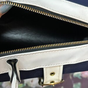 May include: A white and navy blue bag with a gold zipper and a white buckle. The bag is open and the inside is black.
