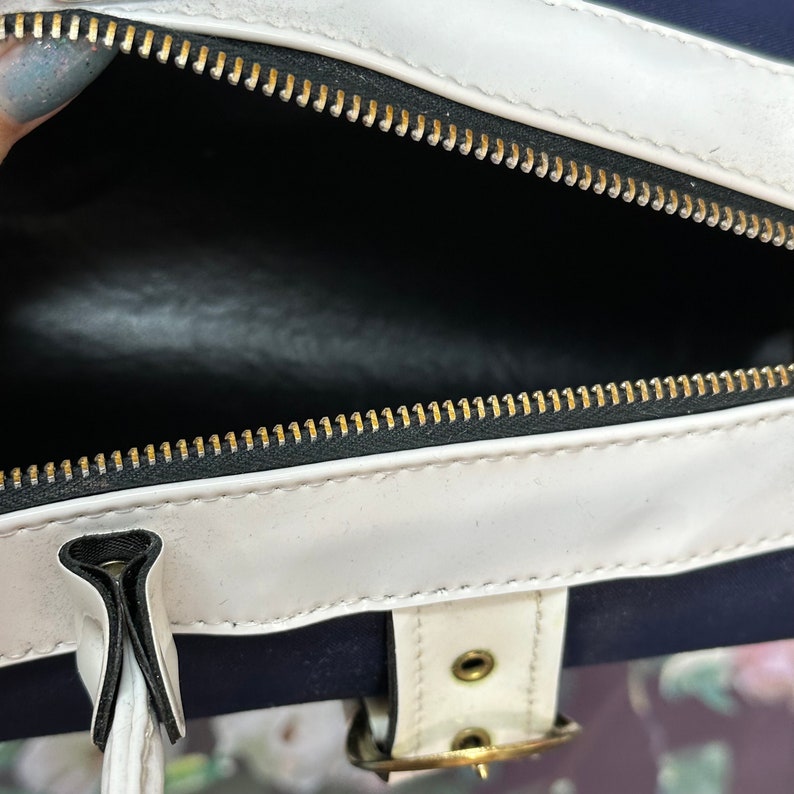 May include: A white and navy blue bag with a gold zipper and a white buckle. The bag is open and the inside is black.