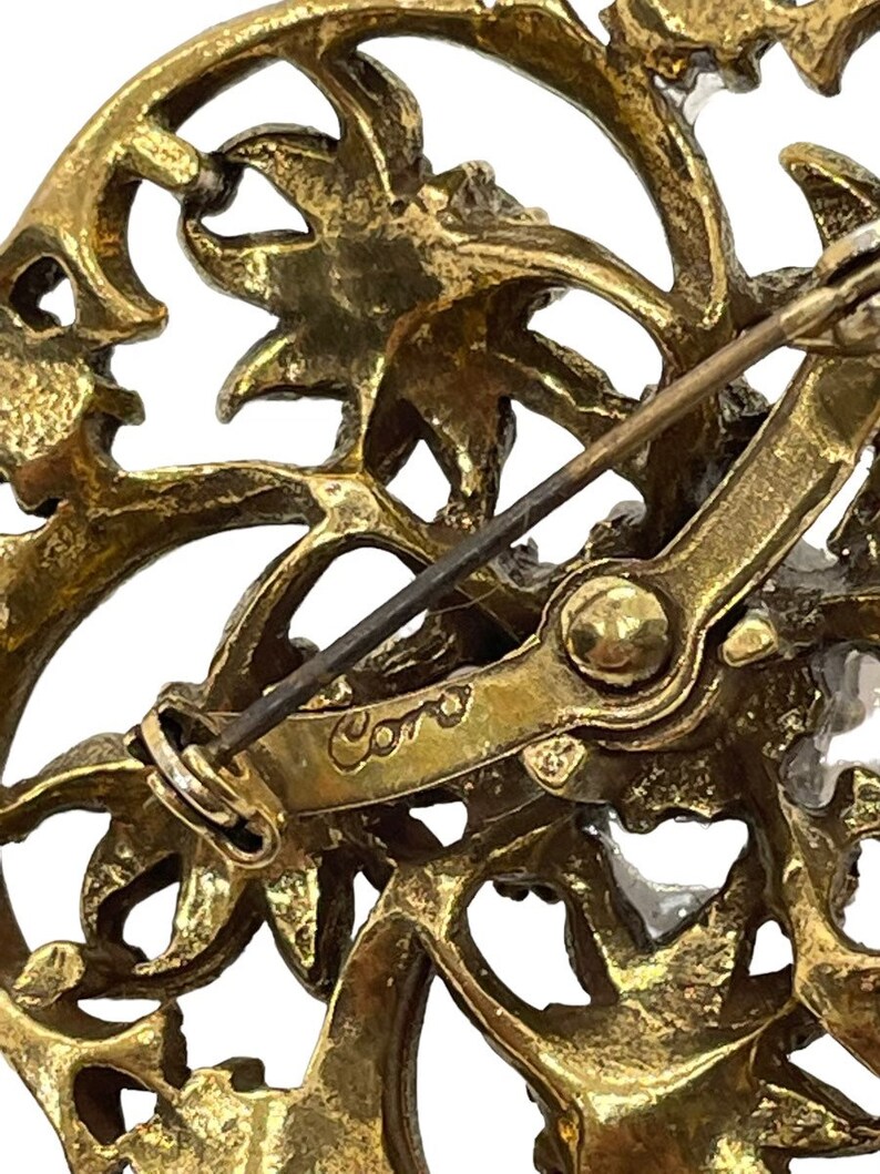 Pu&ograve; includere: Spilla in metallo dorato con un design floreale e una firma Coro sul retro.